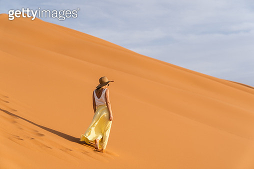A young caucasian woman in a long skirt, shirt and straw hat standing on top of a sand dune and looks towards the sunrise. Sunrise in the Merzouga (Sahara) desert. - gettyimageskorea