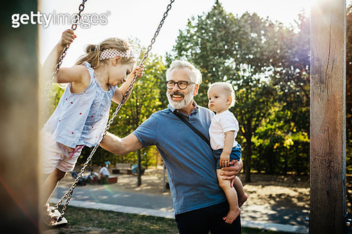 A grandfather having fun, pushing his granddaughter on the swings while holding his grandson on a day out together at the park. - gettyimageskorea